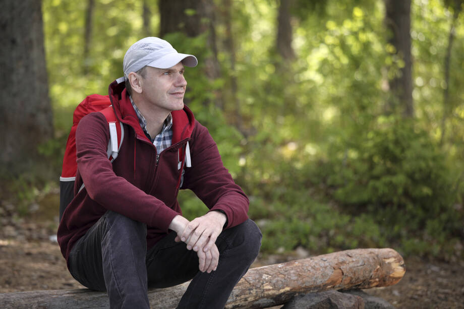 Mies istuu metsässä kaatuneen puun päällä tyytyväisesti hymyillen. 