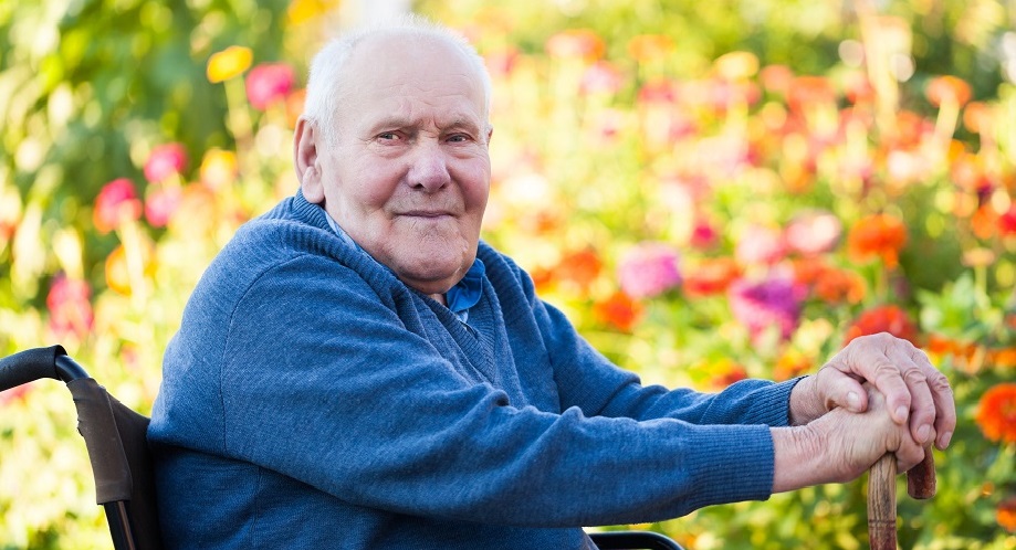 Pyörätuolissa istuva iäkäs mies katsoo kameraa kohti. Taustalla eri värisiä kukkia.