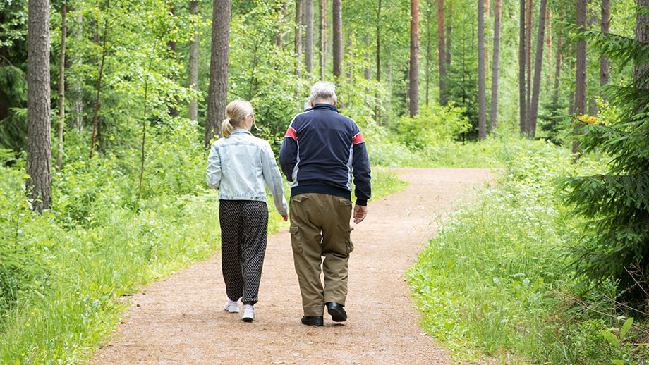 Iäkäs kävelee nuoremman henkilön kanssa kesäisellä metsätiellä.