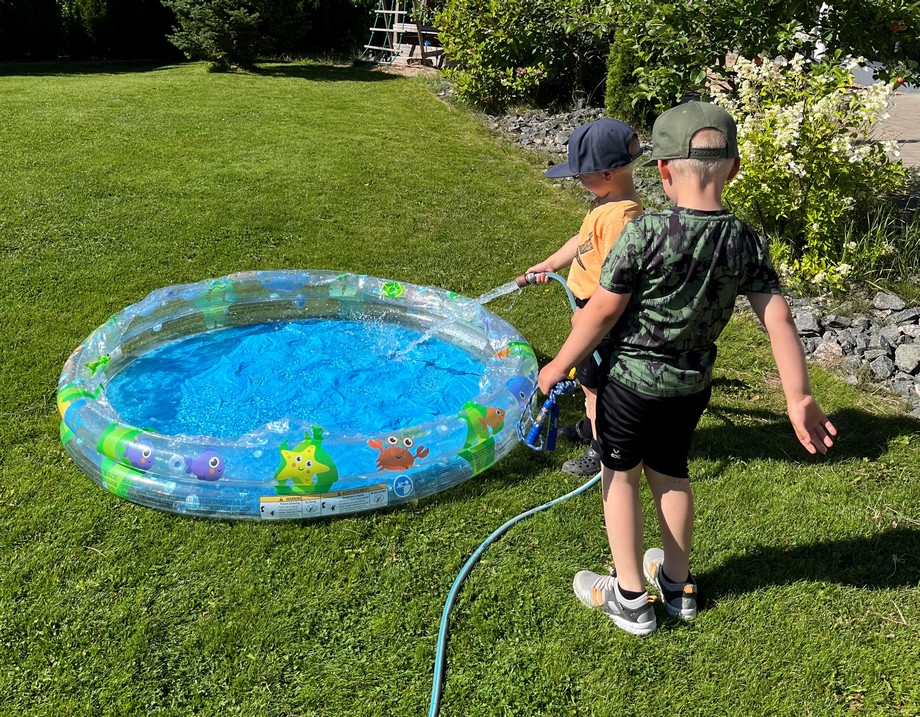 Lippalakkiäiset pikkupojat täyttävät puutarhaletkulla lasten uima-allasta.