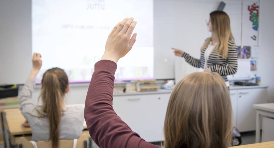 opetaja luokan edessä ja kaksi tyttöä jotka viittaavat. 