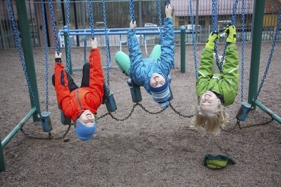 Kolme lasta roikkuu pää alaspäin kiikuissa koulun pihalla.