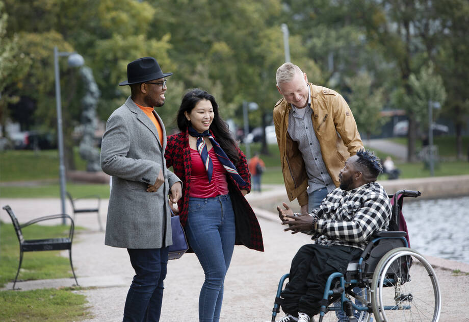 Neljä ihmistä juttelevat kadulla, kolme heistä seisoo ja yksi istuu pyörätuolissa.