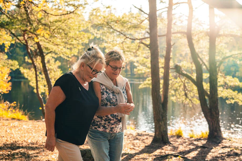 Kaksi ikääntyvää naista kävelee syksyisen lammen rannalla.