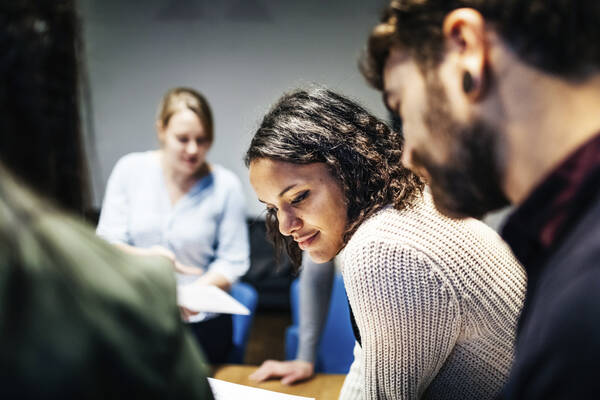 Aikuisia osallistumassa yhteiseen työskentelyyn, etualalla kaksi henkilöä, joista toisella meikatut silmät, ruskea polkkatukka ja vaalea neulepaita ja toisella tumma paita, tummanruskeat hiukset ja parta ja musta korvakoru.