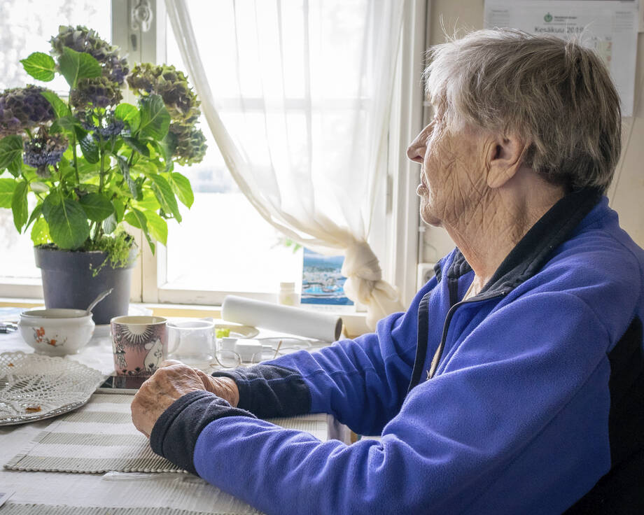 En äldre kvinna sitter vid kaffebordet och tittar ut.