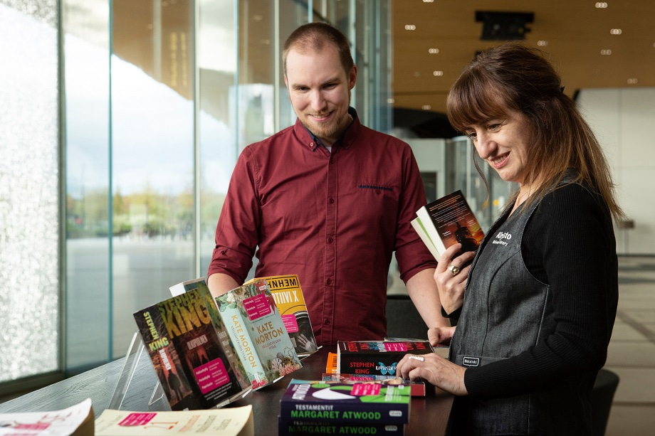 Two smiling persons, a woman and a man, are standing looking at books on a table