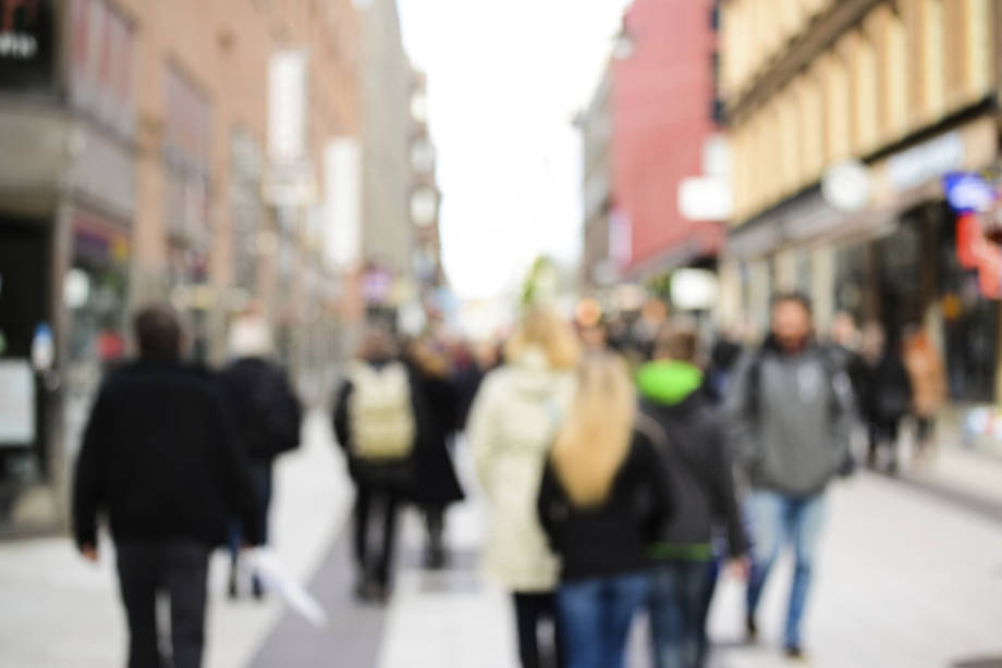 Epätarkka katukuva, jossa ihmiset liikkuvat vilkkaalla ostoskadulla. Kuvan sumennus viittaa väestöön tilastollisena tai tutkittavana kokonaisuutena.
