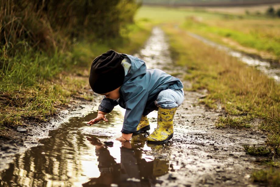 Lapsi leikkii vesilammikossa metsäpolulla sadevaatteissa ja kumisaappaissa. Kuva viestii turvallisesta ja vapaasta ympäristöstä, joka tukee lasten hyvinvointia ja osallisuutta.